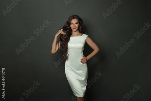 Beautiful smiling woman with long wavy hair wearing white silky dress standing against black studio wall banner background