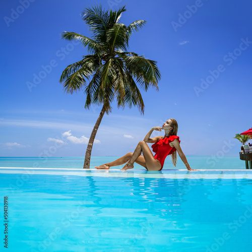 Elegant tanned woman in red swimsuit in pool on tropical Maldives island. Beautiful bikini girl in pool with view on horizon. Sexy model near the pool on beautiful Indian ocean landscape. Travel. 