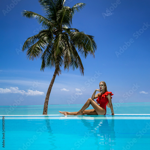 Elegant tanned woman in red swimsuit in pool on tropical Maldives island. Beautiful bikini girl in pool with view on horizon. Sexy model near the pool on beautiful Indian ocean landscape. Travel.  © marcink3333