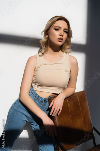 pretty woman in beige top and jeans leaning on chair on white background with shadows.