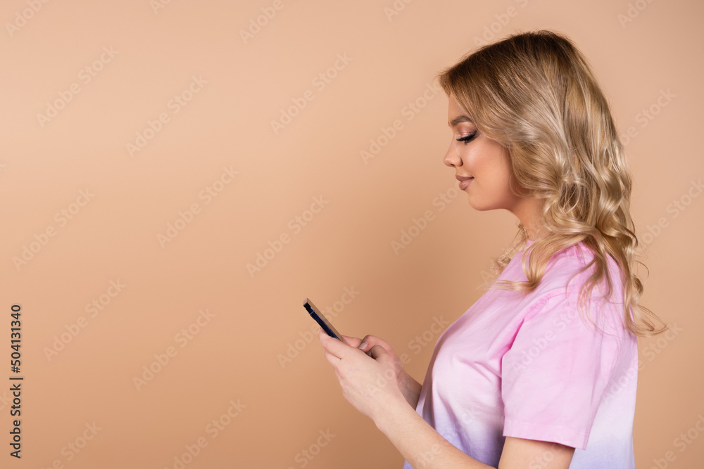 side view of smiling woman with wavy hair messaging on smartphone isolated on beige. side view of smiling woman with wavy hair messaging on smartphone isolated on beige.