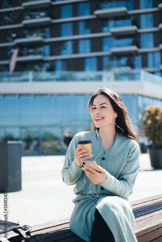 Beautiful business woman enjoying coffee break on free time outdoor with favorite music in earphones, millenial female listening audio book on mobile app