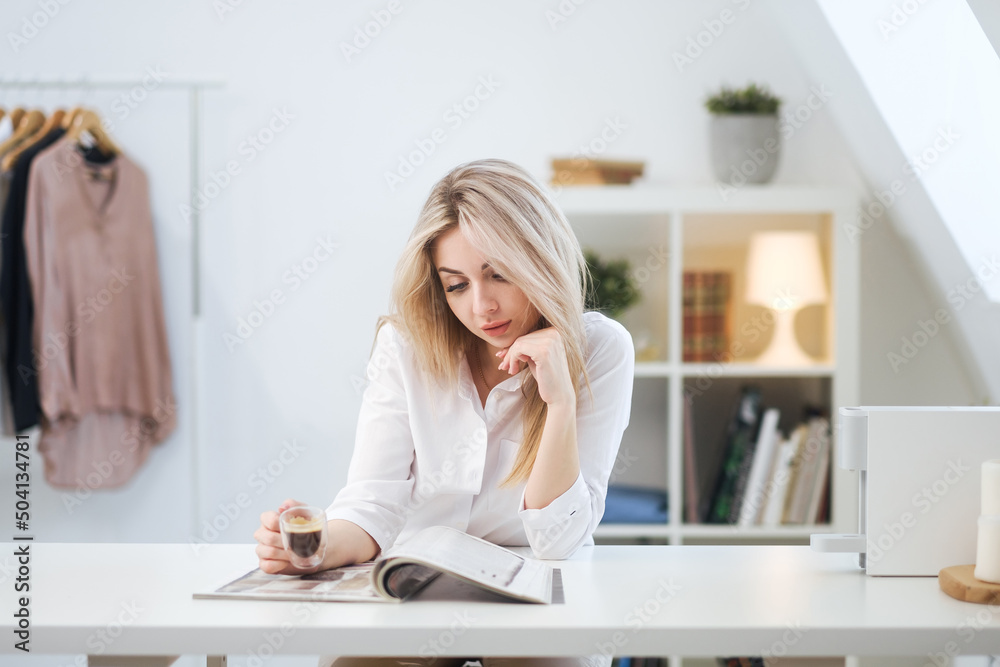 A young beautiful Caucasian blonde woman rests at home. A woman sits at a table, drinking coffee from a glass cup and reading a magazine. A young beautiful Caucasian blonde woman rests at home. A woman sits at a table, drinking coffee from a glass cup and reading a magazine.