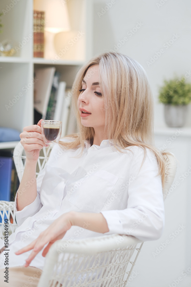 A young beautiful Caucasian blonde woman rests at home. A woman sits at a table, drinking coffee from a glass cup and reading a magazine. A young beautiful Caucasian blonde woman rests at home. A woman sits at a table, drinking coffee from a glass cup and reading a magazine.