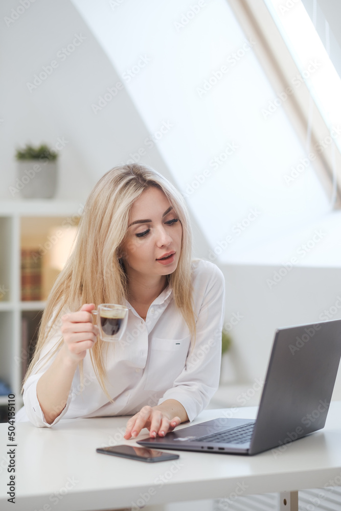Young beautiful caucasian blonde woman working on laptop at home and drinking coffee. Remote work or freelance. Young beautiful caucasian blonde woman working on laptop at home and drinking coffee. Remote work or freelance.