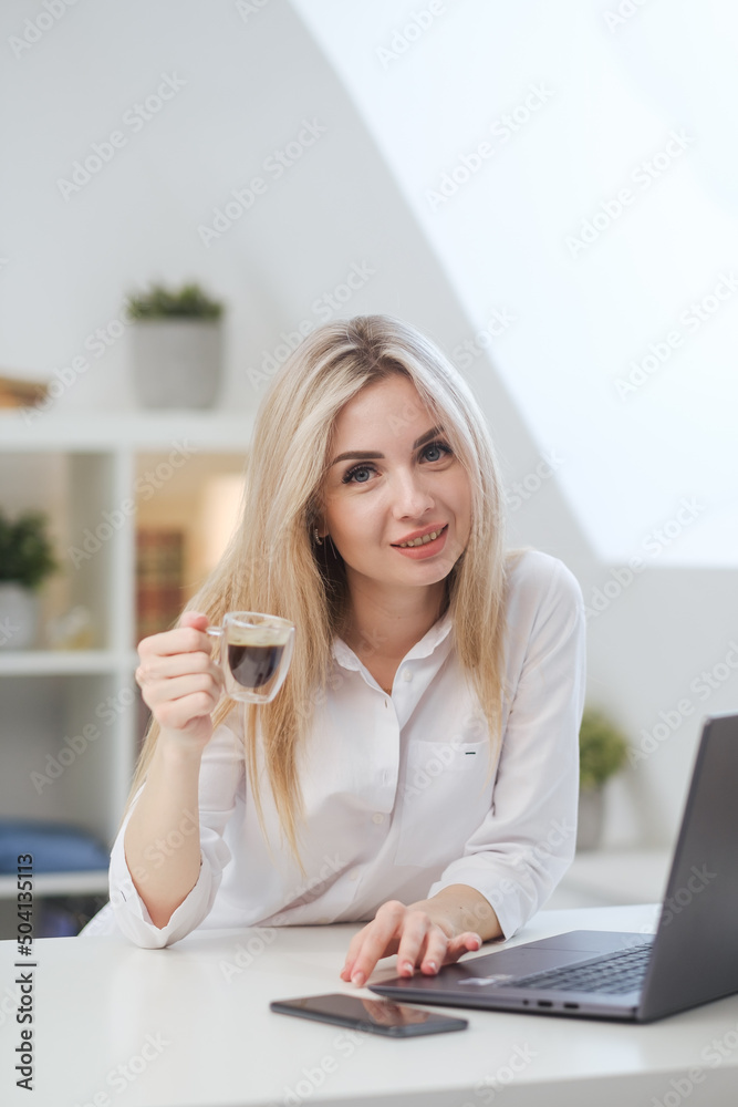 Young beautiful caucasian blonde woman working on laptop at home and drinking coffee. Remote work or freelance. Young beautiful caucasian blonde woman working on laptop at home and drinking coffee. Remote work or freelance.