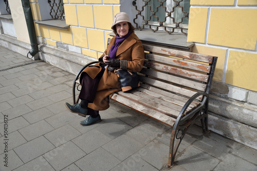 Elderly smiling woman sits on bench in coat and hat in spring city