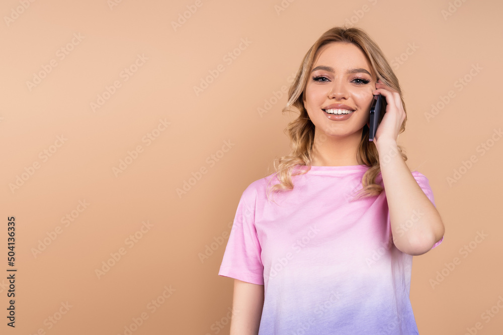 joyful woman in t-shirt talking on smartphone while looking at camera isolated on beige. joyful woman in t-shirt talking on smartphone while looking at camera isolated on beige.
