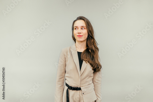 Cute lady in a beige suit stands on a beige background and looks away with a sarcastic smile on her face. Copy space