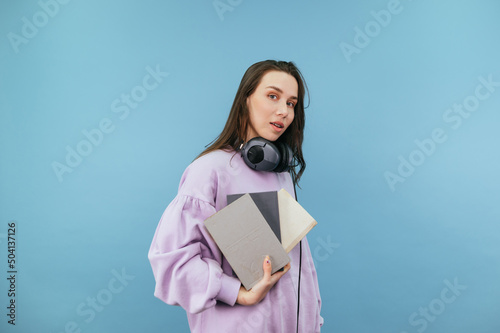Student girl in sweatshirt, headphones and books in her hands stands on a blue background and poses for the camera.