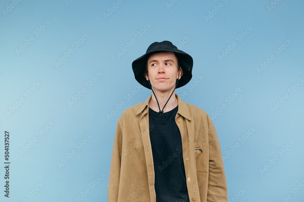 Cute young man in panama and shirt stands on a blue background and looks suspiciously to the side on copy space Cute young man in panama and shirt stands on a blue background and looks suspiciously to the side on copy space