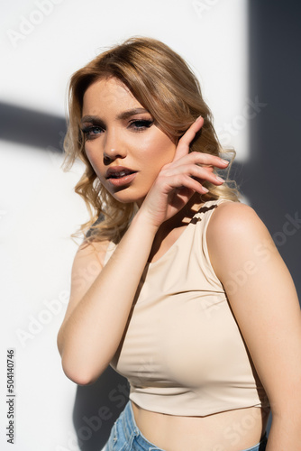 young woman in beige top holding hand near face on white background with shadows.