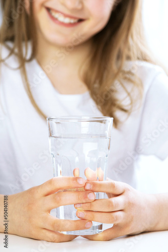 Close-up of a girl in the kitchen drinks clean water from a glass glass. Focus on the glass, face in defocus. The importance of water for children, maintaining water balance