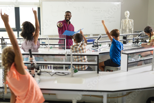 African american young male teacher questioning multiracial elementary students with hands raised