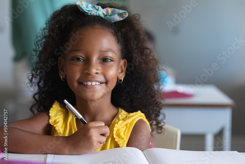 Portrait of smiling african american elementary schoolgirl with curly hair sitting at desk in class