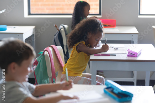 Multiracial elementary school students writing on book while studying at desk in classroom
