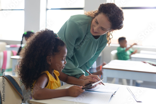 Smiling caucasian young female teacher teaching african american elementary girl at desk