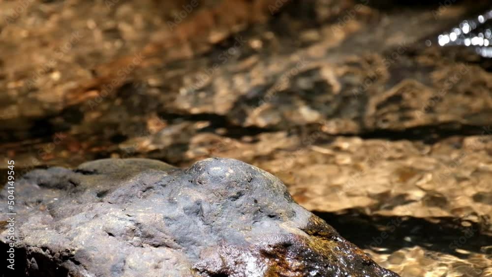 A rock then a stream flowing while ripples reflect sunlight, Kaeng Krachan National Park, Thailand.