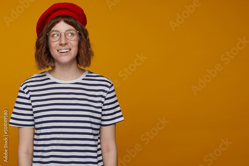 indoor portrait of young ginger female, wears stripped t shirt and glasses posing over orange background looking aside with smile on her face