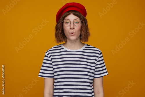 indoor portrait of young ginger female, wears stripped t shirt and glasses posing over orange background looking into camera with surprised facial expression