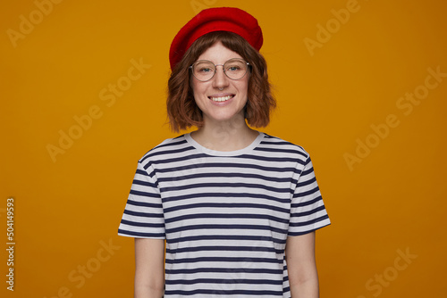 indoor portrait of young ginger female, wears stripped t shirt and glasses posing over orange background looking into camera with broad smile on her face