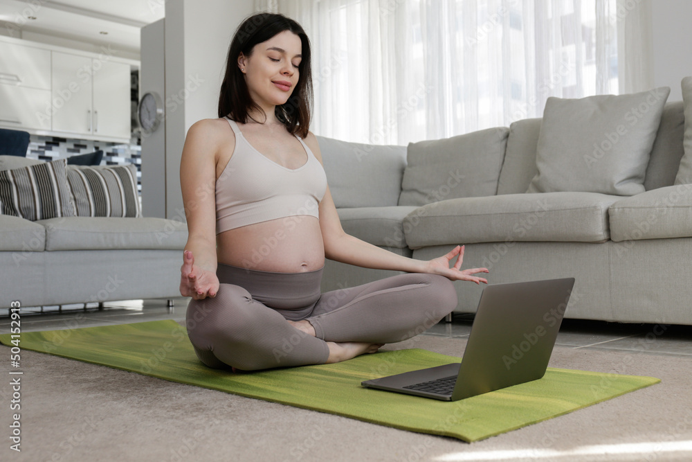 Young beautiful woman on second trimester of pregnancy taking yoga classes online. Pregnant female in sitting on a mat with laptop. Expecting a child concept. Background, copy space, close up. Young beautiful woman on second trimester of pregnancy taking yoga classes online. Pregnant female in sitting on a mat with laptop. Expecting a child concept. Background, copy space, close up.
