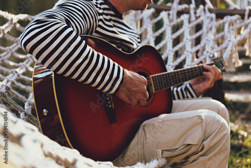 Relaxed young man wearing hipster outfit playing the guitar enjoying the alone time in solitude, lying in a net hammock on beautiful sunny day. Slow living concept. Close up, copy space background.