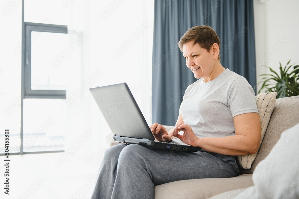 Happy excited grey haired mature woman celebrating online win, using laptop, looking at screen, sitting on couch at home, middle aged female feeling amazed, surprised by unbelievable good news. Happy excited grey haired mature woman celebrating online win, using laptop, looking at screen, sitting on couch at home, middle aged female feeling amazed, surprised by unbelievable good news.