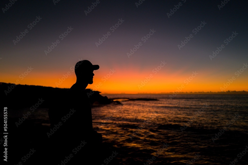 Sillhouette of man wearing baseball cap looking out to the ocean at sunrise