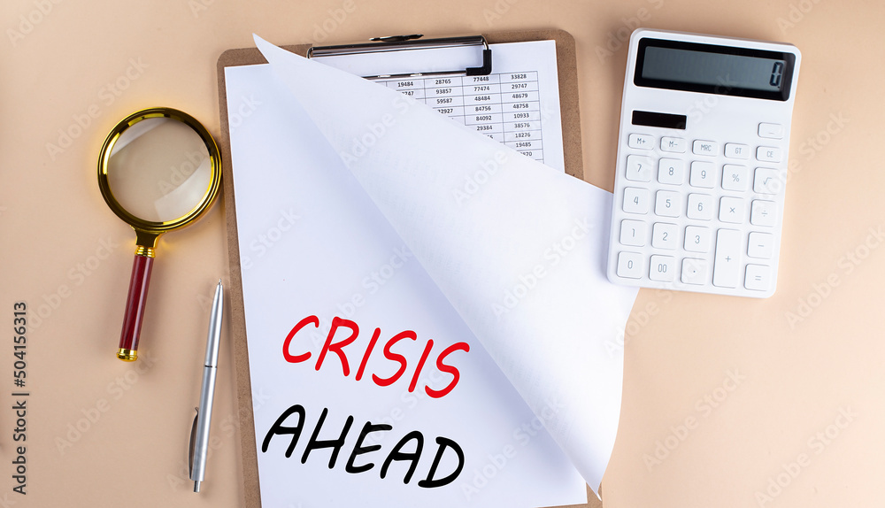 Clipboard with chart and text CRISIS AHEAD with magnifier ,calculator on beige background Clipboard with chart and text CRISIS AHEAD with magnifier ,calculator on beige background