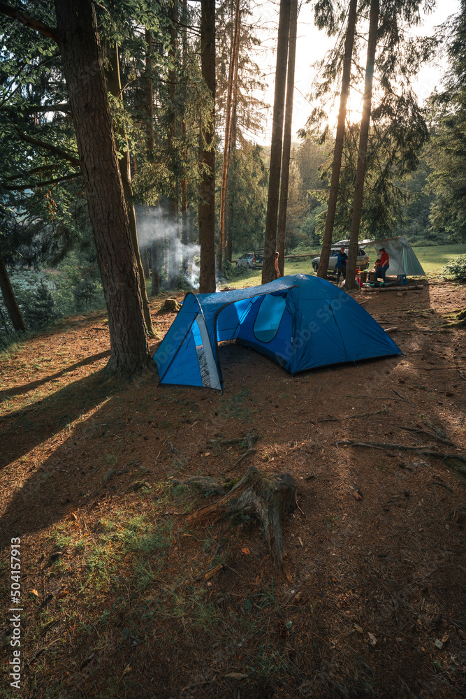 Big dome and camping tent with vestibule. Warm sun rays. Tourist camp among tall pines. Smoke from the fire. Tent. lake shore among the mountains. Vertical photo.