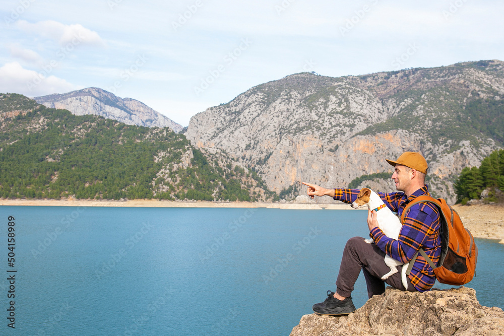 A man and a jack russell terrier enjoying the scenic view of mountain lake. Traveling with the dog concept. Hipster guy backpacking with his pet pup. Background, copy space.