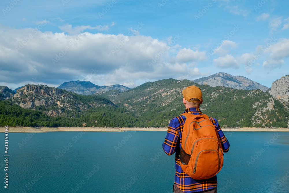 Male traveler wearing flannel shirt and a backpack enjoying beautiful view of a mountain lake. Man experiencing spiritual uplifting from being alone in nature. Scenic background, copy space, back view
