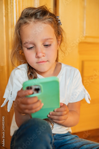 Little girl, blond hair, sit on floor in living room, holds mobile phone in her hands and looks at smartfone photo