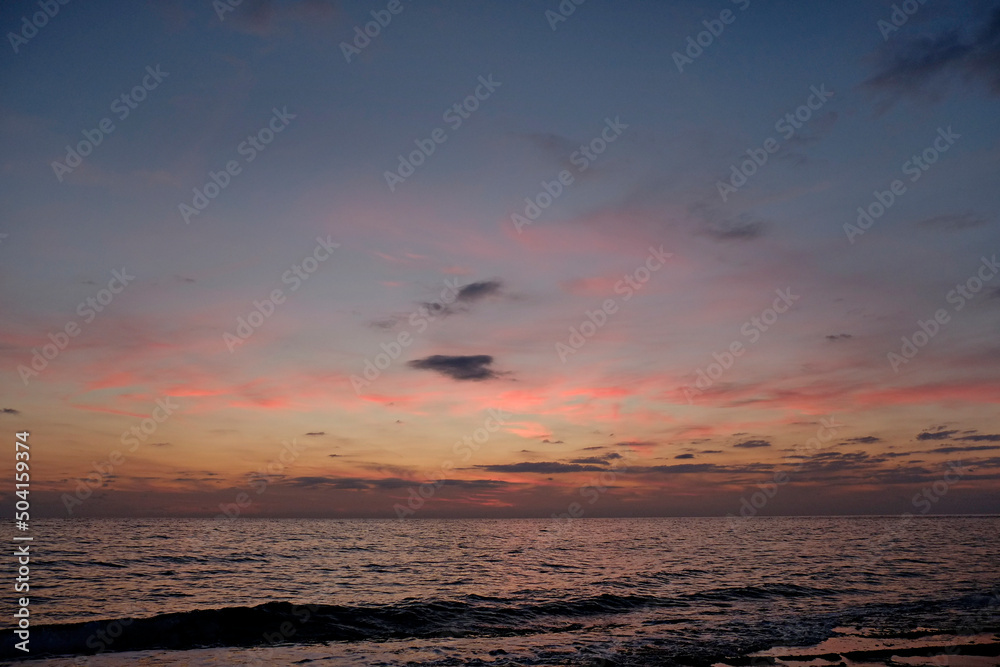 Fototapeta premium Beautiful seascape of sun setting over calm water with wind patterns and colorful pink spindrift clouds in the sky. Ocean view with horizon, background with a lot of copy space for text.
