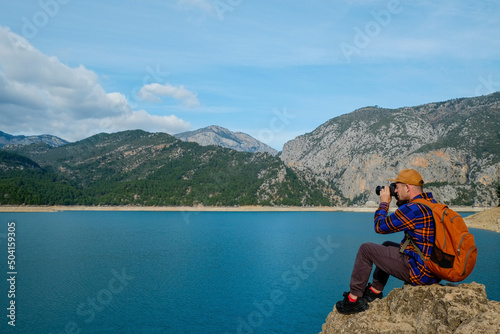 Nature photographer taking photos of beautiful mountain lake on mirrorless camera. Male travel blogger sitting on the edge of a rock capturing a moment on camera. Background, close up, copy space © Evrymmnt