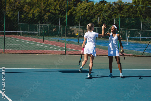 Happy multiracial female doubles team giving high-five while playing tennis at court on sunny day