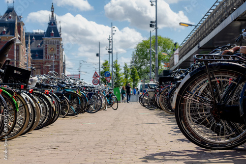 Photography Selective focus of back wheel of bicycle under blue sky, Outdoor public parking area with blurred view of Amsterdam central station building, Cycling is a common mode of transport in the Netherlands