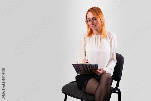 A girl, in a white blouse, sits on a chair with a folder in her hands, on a gray background
