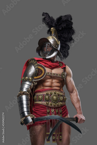 Shot of muscular ancient gladiator dressed in red cape with armor and helmet holding two swords.