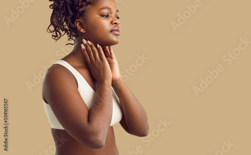 Woman suffers from throat pain due to inflamed tonsil glands or lymphadenitis lymph node infection. Beautiful black lady touches painful neck area isolated on solid beige color text space background photo