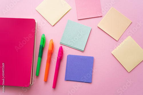 top view of colorful pens near bright notebook and blank paper notes on pink.