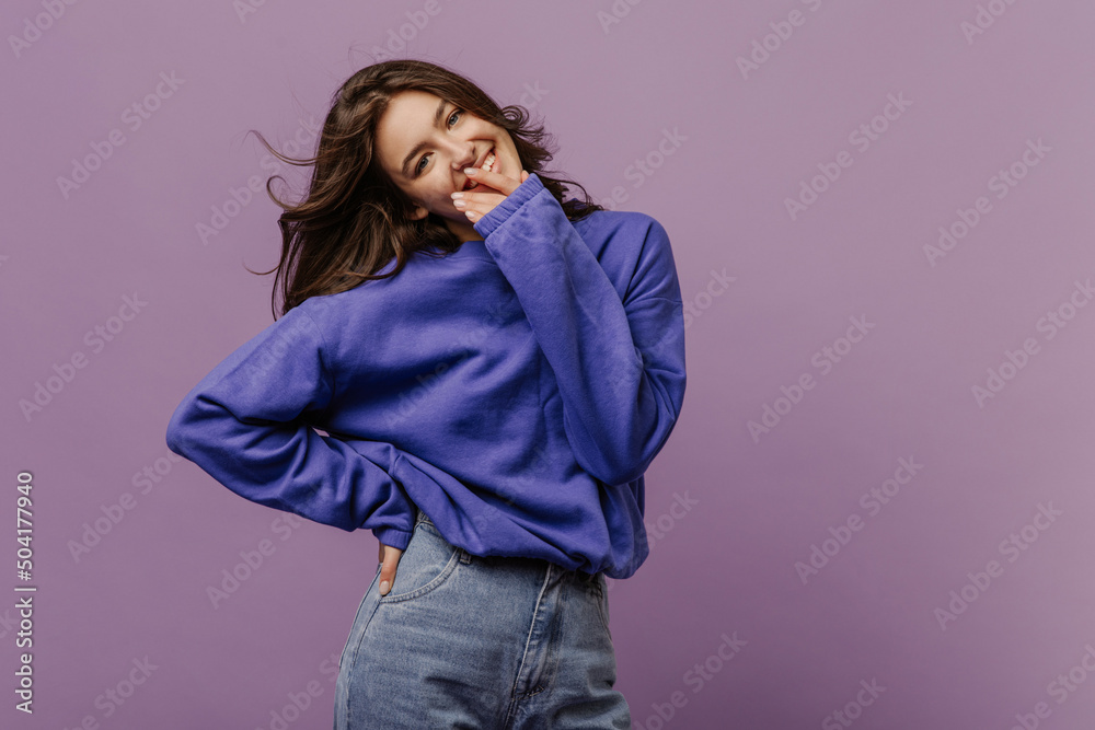 Smiling young caucasian woman smiling looking at camera holding hand near face on purple background. Dark-haired beauty wears blue sweatshirt and jeans. Emotion concept Smiling young caucasian woman smiling looking at camera holding hand near face on purple background. Dark-haired beauty wears blue sweatshirt and jeans. Emotion concept