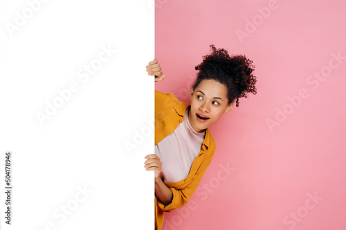 Amazed african american girl with curly hair, peeking out from blank white board with mockup template and copy space for presentation or advertising, stand on pink background, smiles. Mock-up concept photo