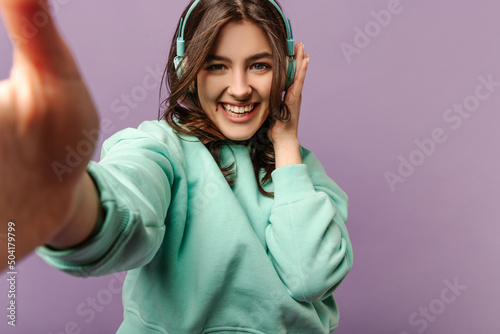 Beautiful caucasian young woman with dark hair wears headphones, takes selfie on purple background. Model in turquoise hoodie enjoys listening to music.