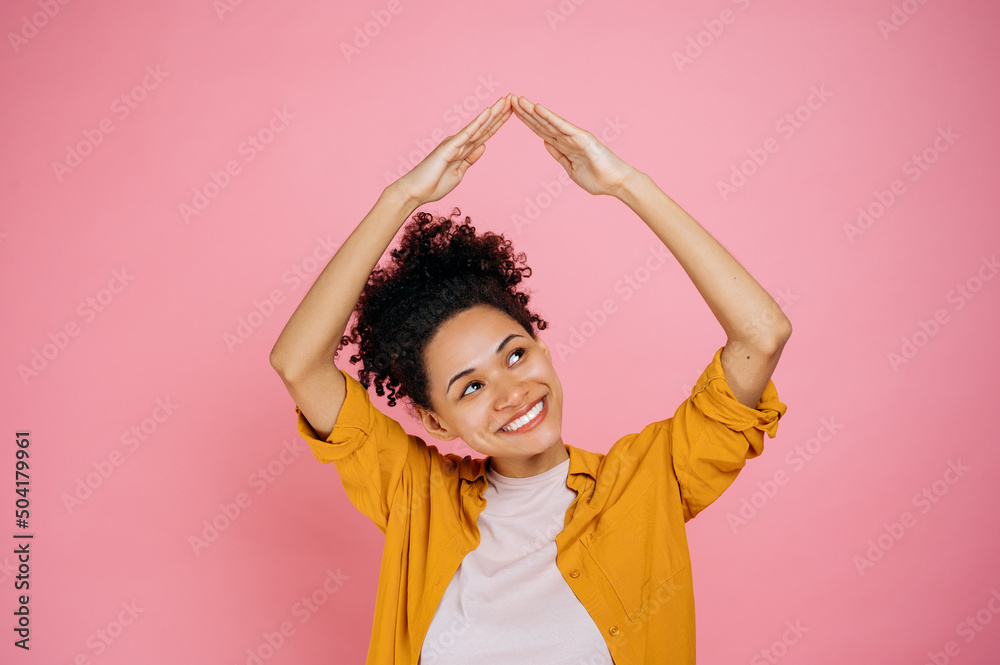Cheerful pretty, african american curly haired girl in casual clothes, is feeling safe under house roof gesture, looks up and smiling, dreaming of home, standing on isolated pink background Cheerful pretty, african american curly haired girl in casual clothes, is feeling safe under house roof gesture, looks up and smiling, dreaming of home, standing on isolated pink background