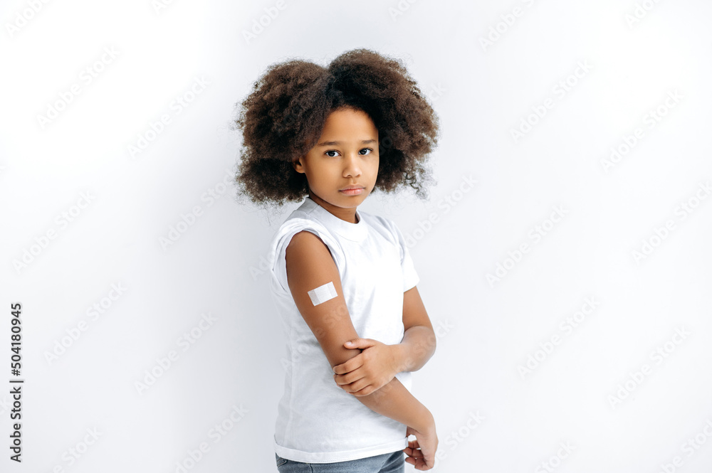 Upset African American curly haired little girl, with a band-aid on her shoulder after vaccination, stand on isolated white background, looks at camera. Virus protection, immunity concept Upset African American curly haired little girl, with a band-aid on her shoulder after vaccination, stand on isolated white background, looks at camera. Virus protection, immunity concept