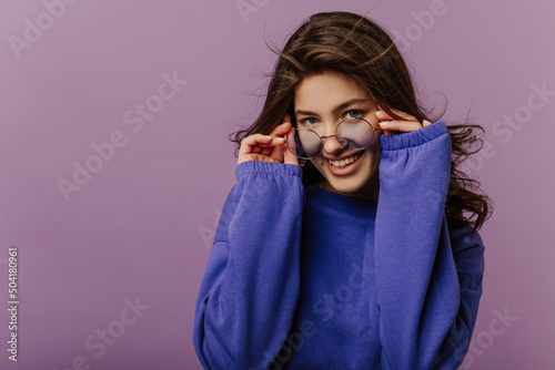 Cool young caucasian girl looks at camera with her glasses down on nose standing on purple background. Brunette with wavy hair wears blue sweatshirt. Leisure lifestyle and beauty concept.