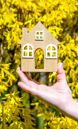 The girl holds the house symbol against the background of blossoming forsythia 