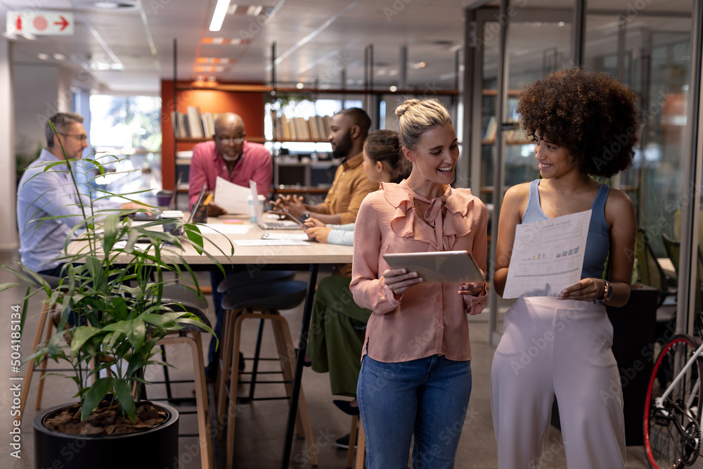 Obraz premium Smiling biracial businesswomen discussing together against colleagues working at modern workplace